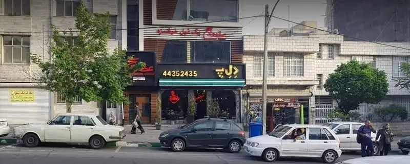 نمایی از فست فود د ل پ در تهران، تابلوی رستوران با شماره تماس 44352435، خودروها و مردم در حال عبور در یک روز آفتابی.