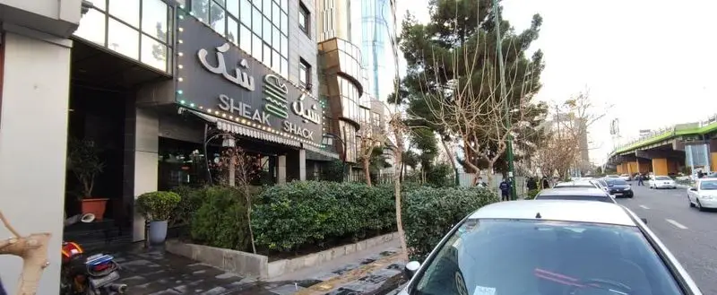 نمای بیرونی جذاب رستوران شیک شک (Sheak Shack) با تابلوی روشن، لوگوی برگر، فضای سبز شهری و خودروهای پارک شده در کنار خیابان.