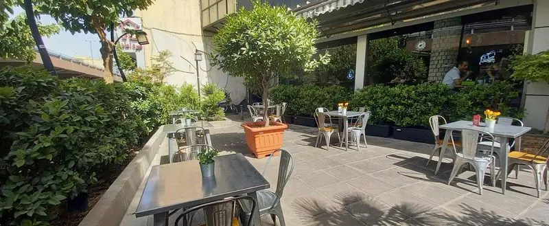 نمای خارجی کافه رستوران شیک شک: فضای باز دلپذیر با میز و صندلی‌های فلزی، درختان در گلدان و بوته‌های سبز در اطراف.