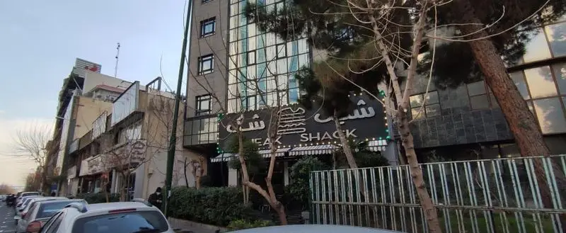 تصویری از نمای بیرونی رستوران شیک شک (Steak Shack) با تابلوی روشن و درختان در جلوی آن، در یک خیابان شلوغ تهران.