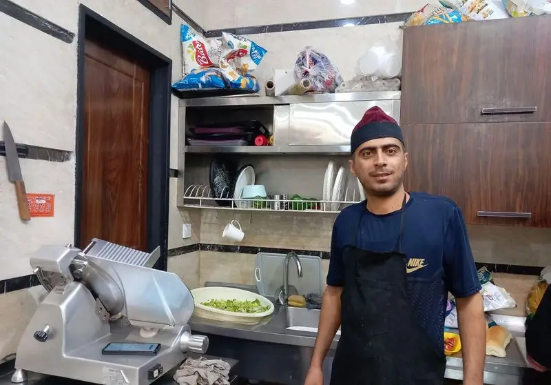 آشپز فستفود گل رز در آشپزخانه مدرن و بهداشتی، مشغول آماده‌سازی ساندویچ و سالاد با دستگاه اسلایسر و مواد تازه.