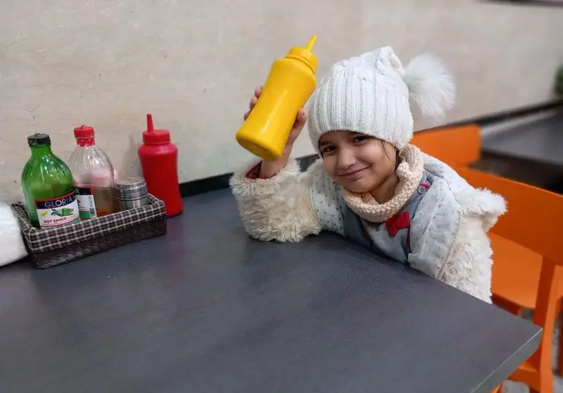 کودکی شاد و خندان در فستفود گل رز، با کلاه زمستانی سفید و سس خردل زرد در دست، پشت میزی پر از چاشنی‌های متنوع؛ لحظاتی دلنشین.