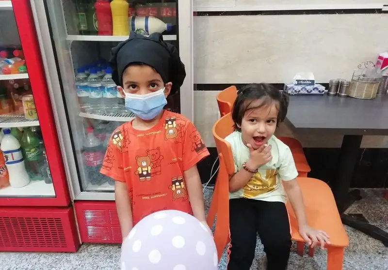 کودکان شاد در فستفود گل رز، پسربچه با کلاه سرآشپز و ماسک کنار یخچال نوشیدنی و دختربچه با نشاط در محیط دوستانه رستوران