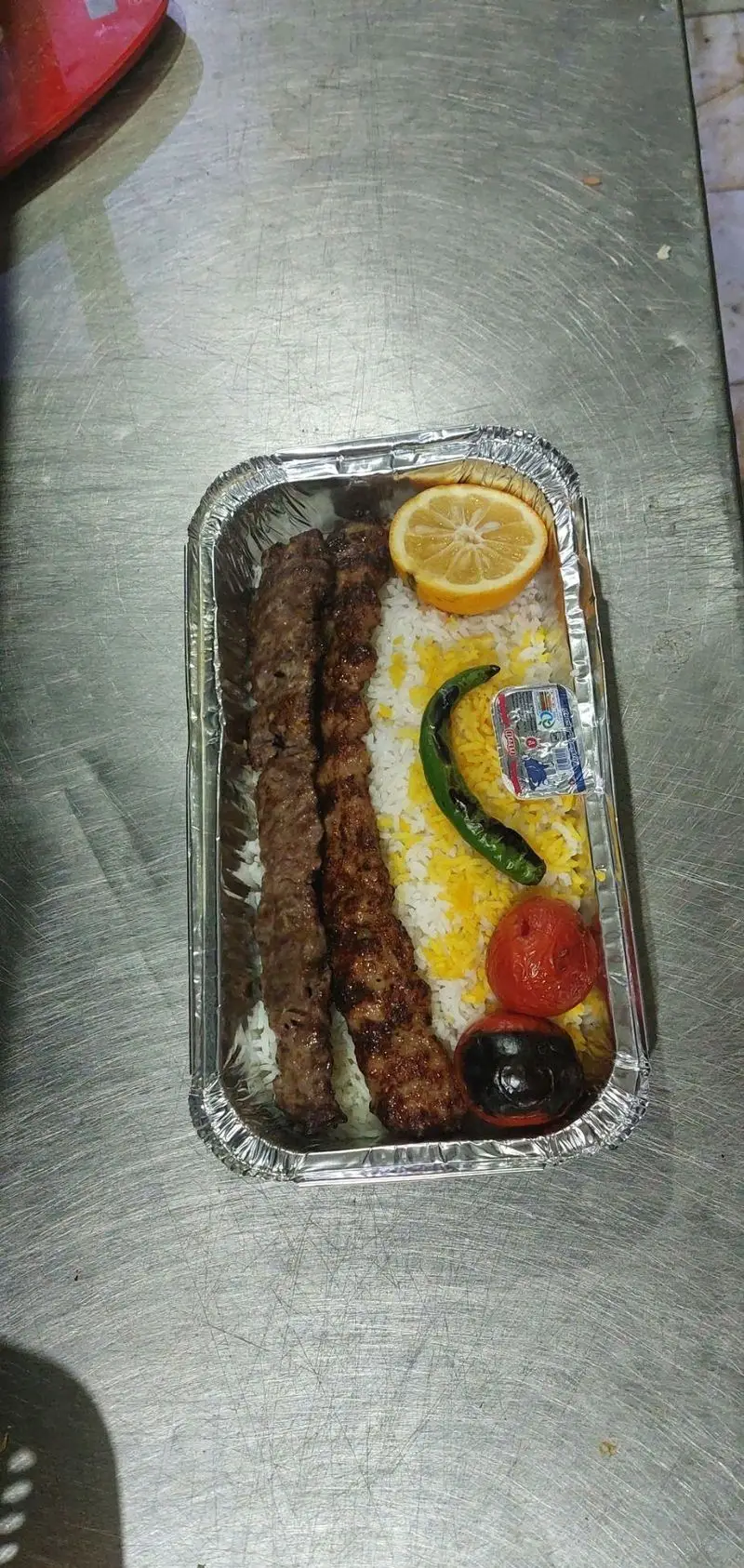 غذای ته چین تهرانپارس: چلوکباب کوبیده لذیذ و اصیل ایرانی در ظرف بیرون بر آلومینیومی، همراه با برنج زعفرانی، گوجه کبابی، لیمو و فلفل.