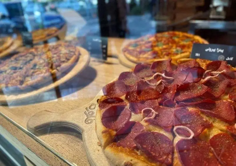 پیتزاهای متنوع کافه فلوره در ویترین شیشه‌ای، از جمله پیتزا پپرونی پرملات با پنیر و پیاز، آماده سفارش و لذت بردن.