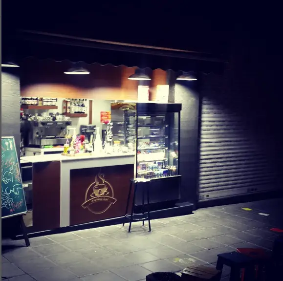 ورودی دنج کافه پرشین در شب با نورپردازی گرم، شامل میز سفارش، تجهیزات قهوه‌ساز، ویترین جذاب و صندلی‌های فضای باز برای مشتریان.