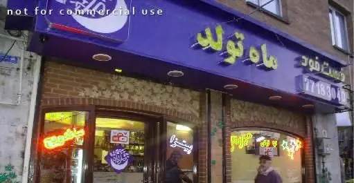 نمای بیرونی فست فود ماه تولد با تابلوی آبی رنگ و حروف زرد درخشان، به همراه ویترین شیشه‌ای و چراغ‌های نئون رنگارنگ.