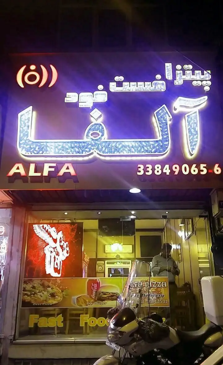نمای شب از سردر فست فود آلفا با تابلوی نئون درخشان، ارائه دهنده پیتزا، برگر و فست فود با تحویل رایگان و شماره تماس.