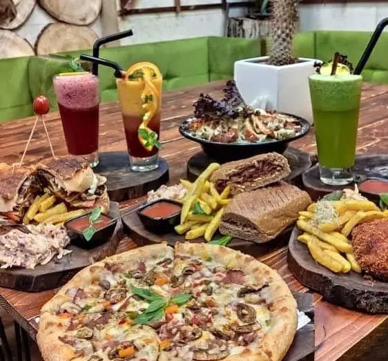 تصویری از میز شلوغ و متنوع کافه چوب سبز با پیتزاهای داغ، ساندویچ و برگرهای تازه، سالاد و آبمیوه‌های طبیعی و رنگارنگ.