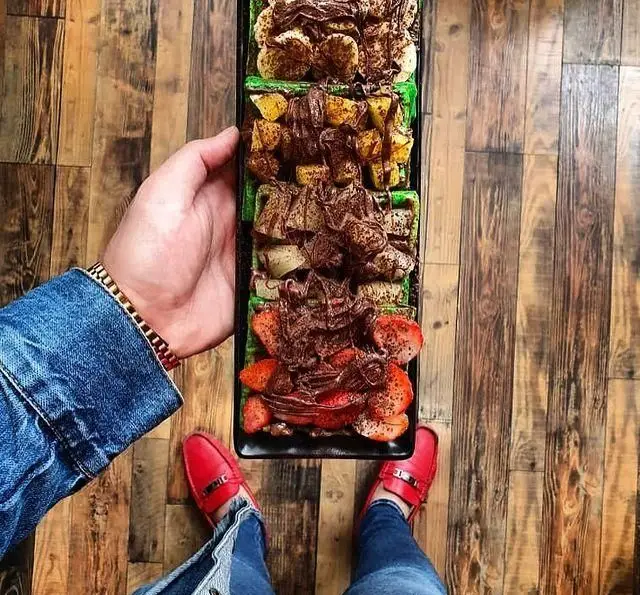 سینی جذاب دسرهای کافه چوب سبز، وافل یا کرپ شکلاتی با موز، توت فرنگی و میوه‌های دیگر در دست مشتری روی کفپوش چوبی.