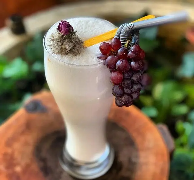 نوشیدنی میوه‌ای سفیدرنگ و شیک کافه چوب سبز، مزین به خوشه‌ی انگور قرمز آبدار، نی فلزی خاص و غنچه رز خشک شده در فضایی دنج.
