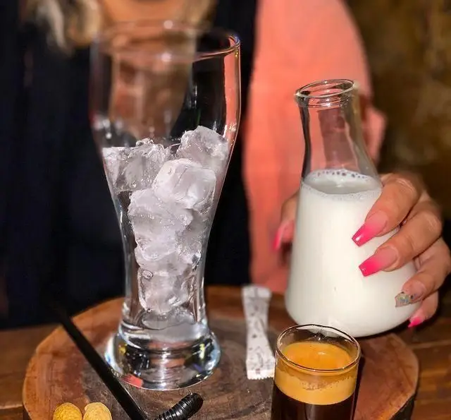 کافه چوب سبز: آماده‌سازی نوشیدنی قهوه خنک، شامل لیوانی پر از یخ، شیشه شیر و شات اسپرسو روی سینی چوبی، محیطی دلپذیر.