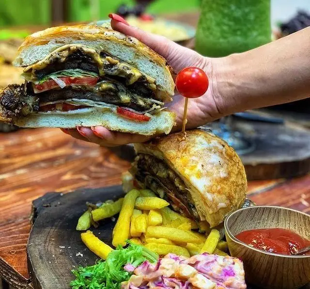 تصویر برگر ویژه کافه چوب سبز با پنیر و گوجه، همراه سیب‌زمینی سرخ‌کرده، کلم‌سالاد، سس گوجه و نوشیدنی سبز.