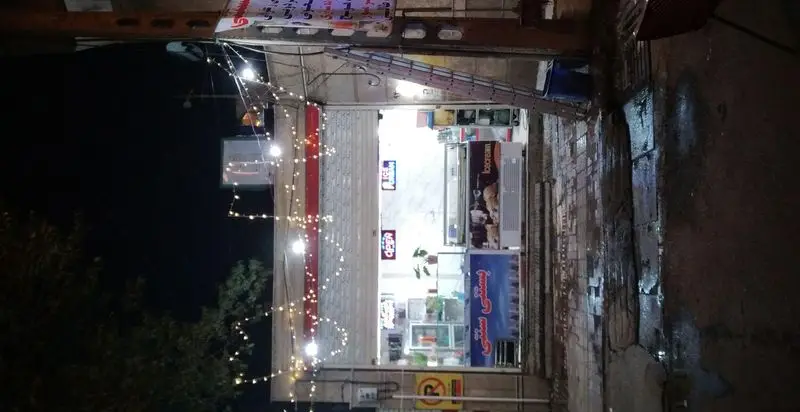 نمای بیرونی شبانه آبمیوه و بستنی جانیار با نورپردازی ریسه ای و تابلوهای روشن، آماده پذیرایی از مشتریان با انواع دسرهای خنک.