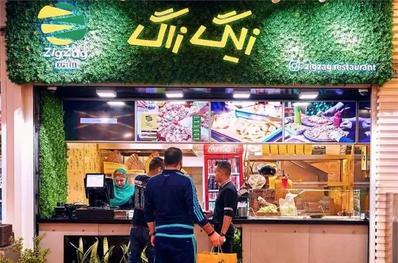 تصویری از نمای بیرونی فست فود و رستوران زیگ زاگ با لوگوی سبز و زرد، نمایش غذاهای متنوع و مشتریان در حال سفارش.