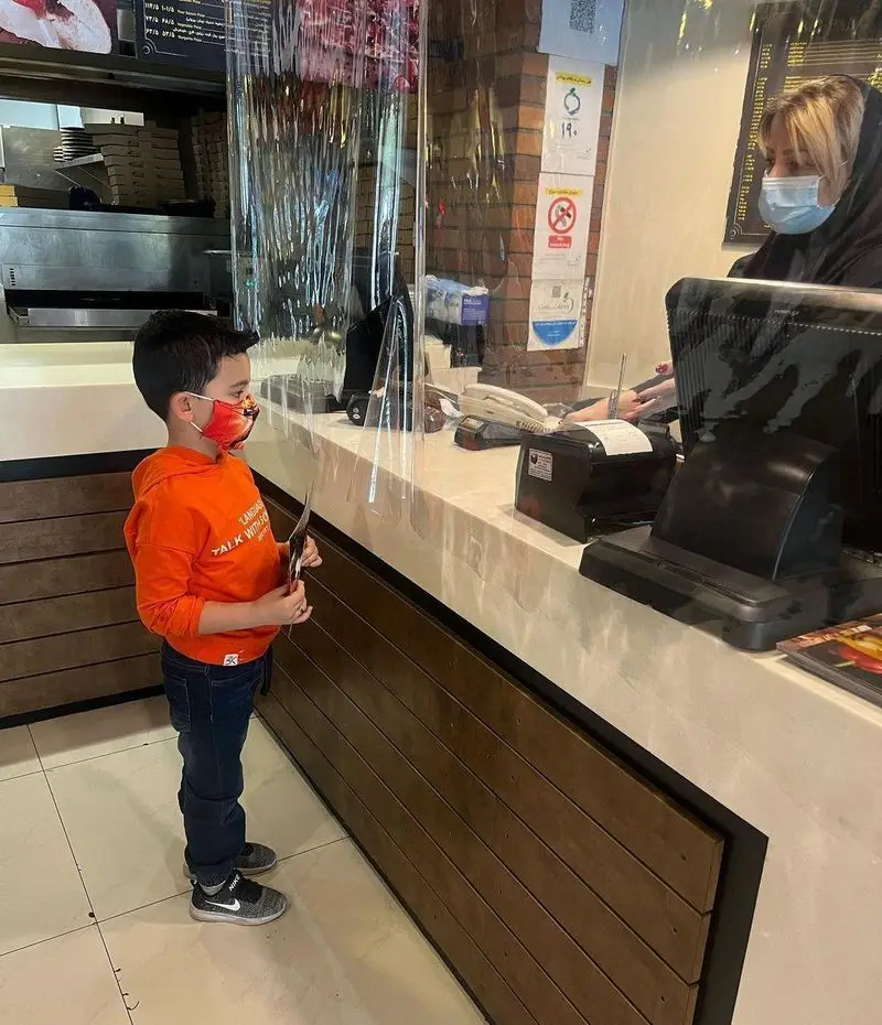 پسر بچه ای با ماسک نارنجی در حال سفارش غذا از پیشخوان رستوران بیگ بنگ، با وجود محافظ پلاستیکی برای امنیت مشتریان.