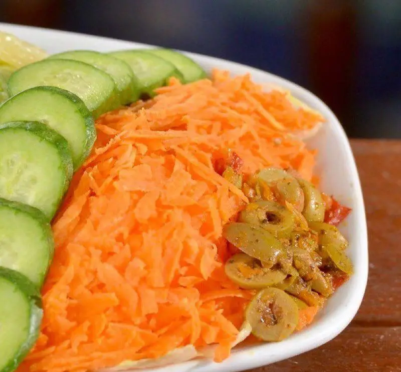 تصویری نزدیک از سالادی رنگارنگ و تازه شامل خیارهای برش خورده، هویج رنده شده و زیتون سبز چاشنی‌دار از رستوران باغ طهرون.