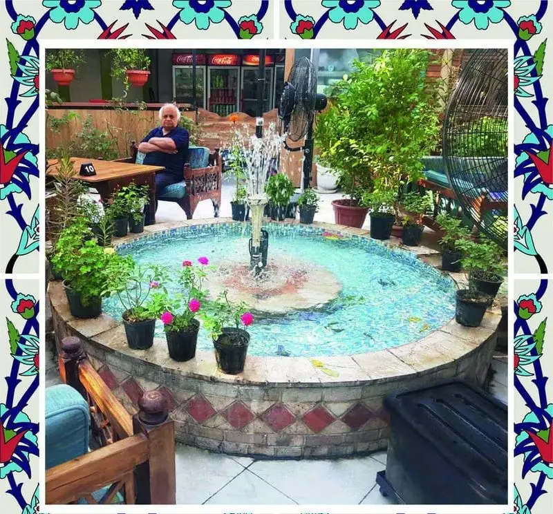 فضای دنج و زیبای رستوران باغ طهرون با فواره مرکزی، گیاهان سبز و گل‌های رنگارنگ. مشتری در حال استراحت در این محیط آرام و دلنشین.