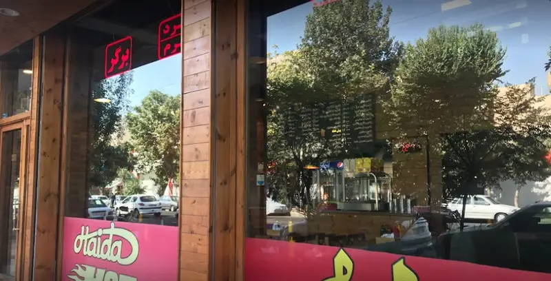 تصویری از نمای بیرونی فست فود هایدا با ویترین شیشه ای و تابلوی برگر روشن. انعکاس درختان، خیابان و ماشین ها در شیشه دیده می شود. لوگوی هایدا در پایین.