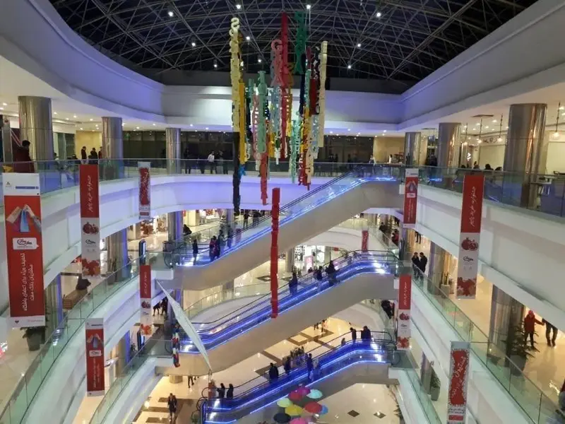تصویر دیدنی از داخل مرکز خرید بزرگ معین مال با پله برقی‌های درخشان آبی، دکوراسیون زیبا و مردمی که مشغول خرید هستند.