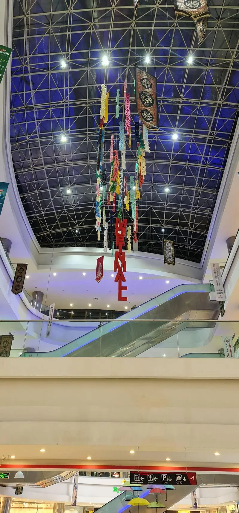 تصویری زیبا از فضای داخلی مرکز خرید معین مال، شامل سقف شیشه‌ای مدرن، لوستر تزئینی بزرگ با حروف رنگی و تابلوهای خوشنویسی اسلامی، و پله‌برقی‌ها.