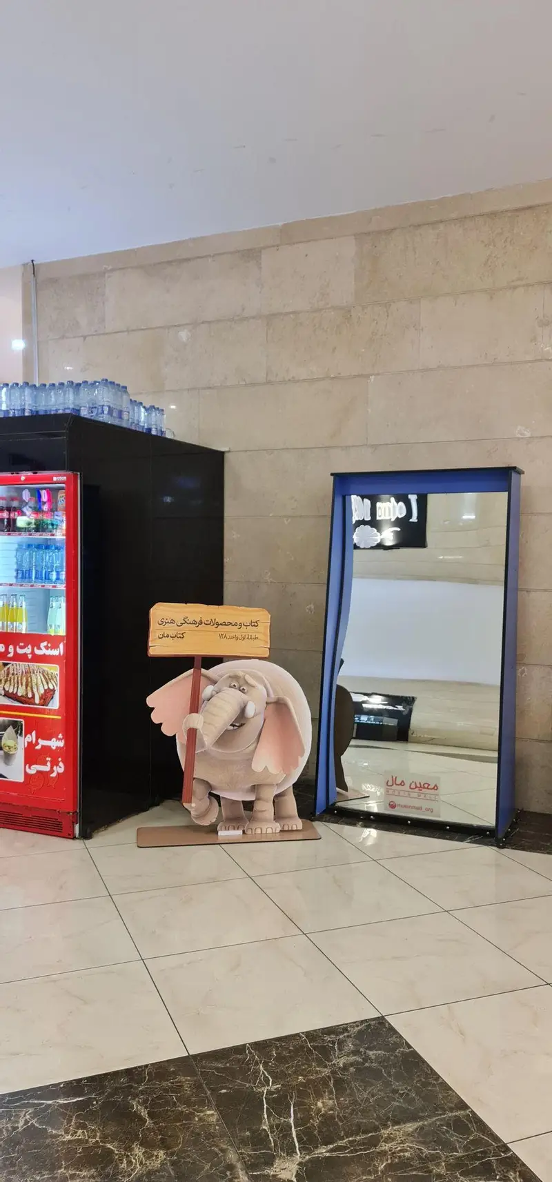 تصویر داخلی از معین مال با استند فیل کارتونی معرفی‌کننده فروشگاه کتاب و محصولات فرهنگی هنری کتاب مان در طبقه اول.