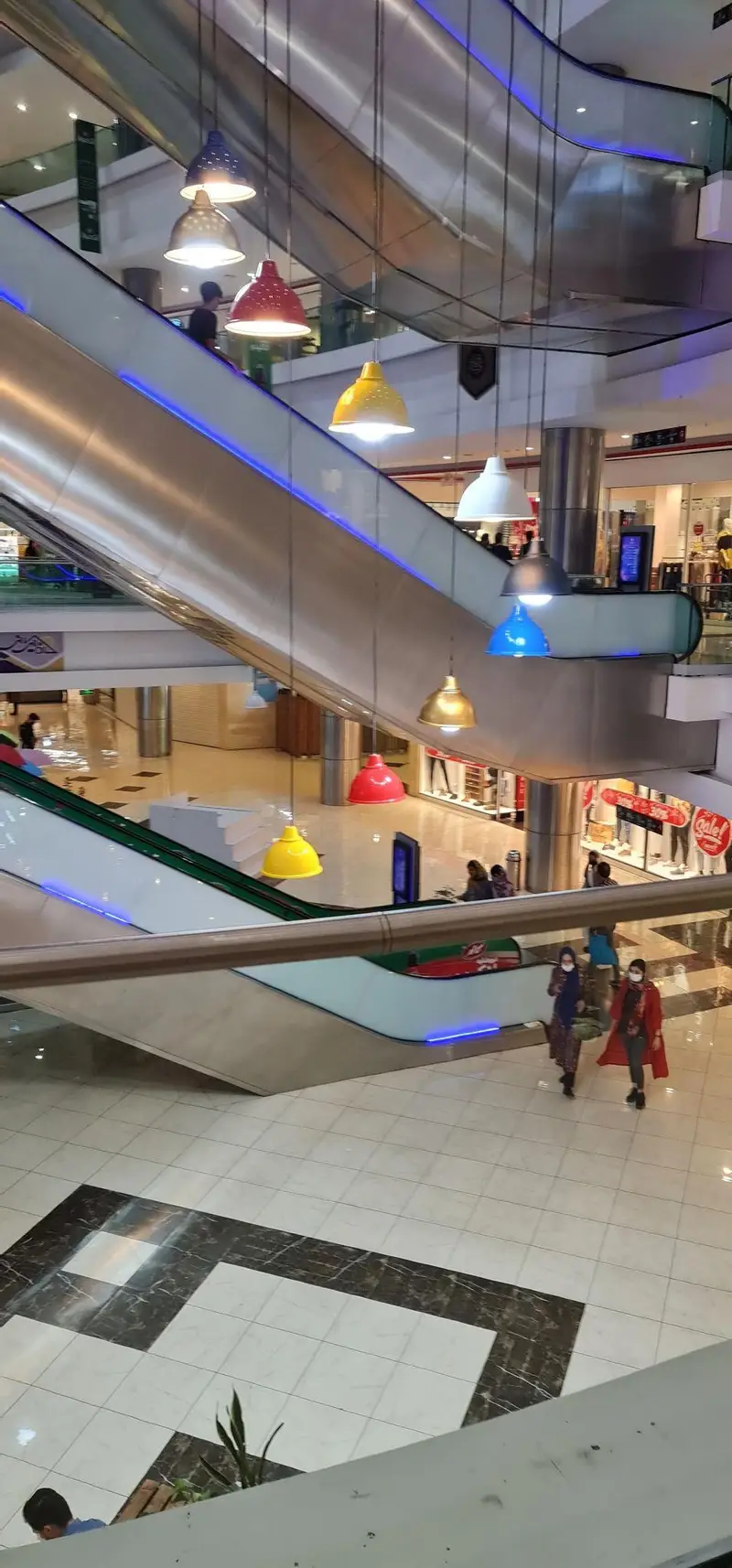 تصویری از طراحی داخلی مدرن معین مال، با پله برقی‌های استیل و چراغ‌های آویز رنگارنگ که فضای پر جنب و جوش مرکز خرید را نشان می‌دهد.
