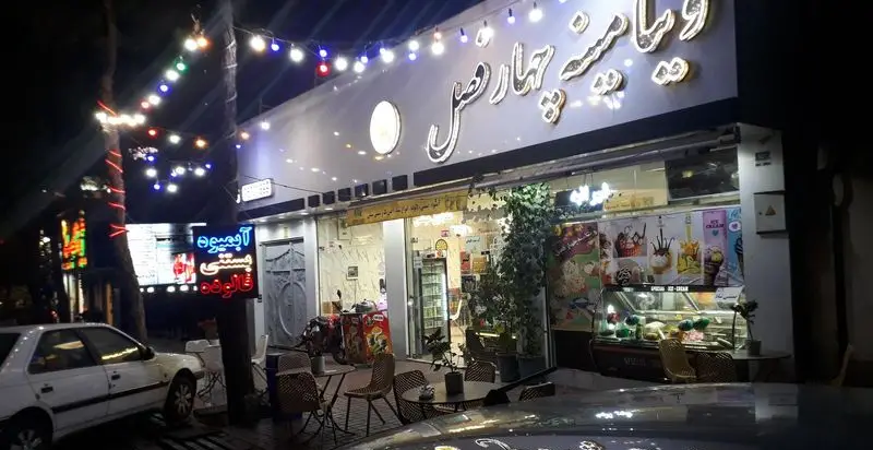 تصویری از نمای شبانه مغازه ویتامینه چهارفصل با چراغ‌های تزئینی، تابلوهای نئونی آبمیوه، بستنی و فالوده، و فضای باز کافه با میز و صندلی.