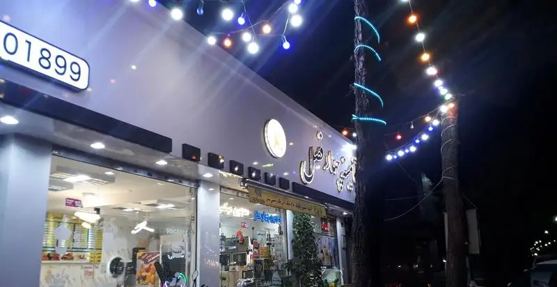 نمای بیرونی آبمیوه فروشی ویتامینه چهارفصل در شب، با تابلو روشن، تزئینات ریسه ای و چراغ‌های رنگارنگ روی ساختمان و درختان.