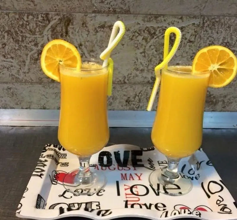 دو لیوان آب پرتقال تازه، خنک و گوارا با تزئین پرتقال و نی‌های رنگی روی سینی زیبا، ایده‌آل برای رستوران بی بی گل.