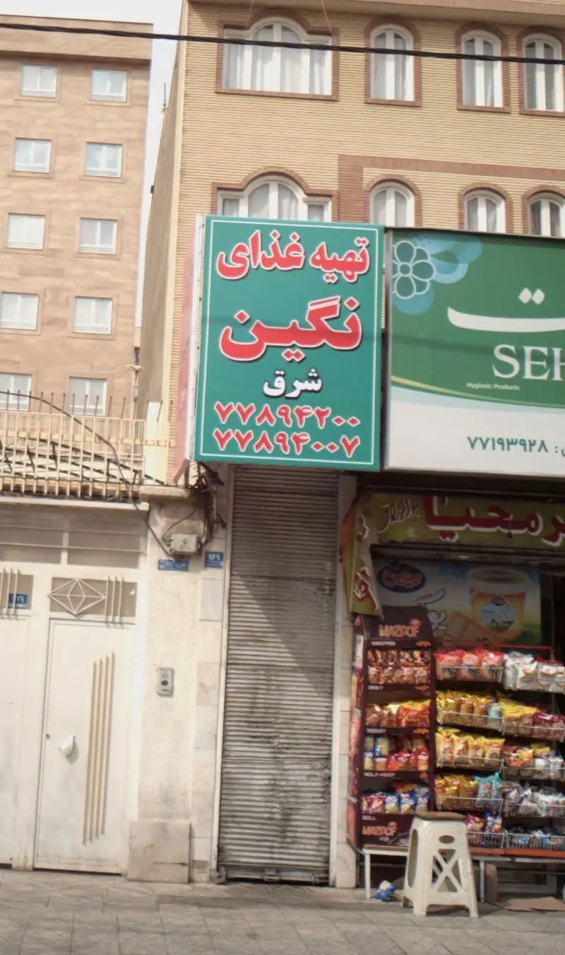 تابلوی سبز رنگ تهیه غذای نگین شرق به همراه شماره تماس، یک رستوران بیرون بر و کترینگ در نمای شهری ایران.