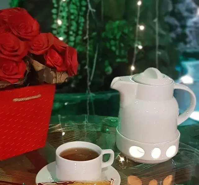 یک فنجان چای گرم کنار قوری سفید با گل‌های کاغذی قرمز، در فضای آرام و زیبای سفره خانه سنتی ماندگار، مناسب لحظات دنج شما.