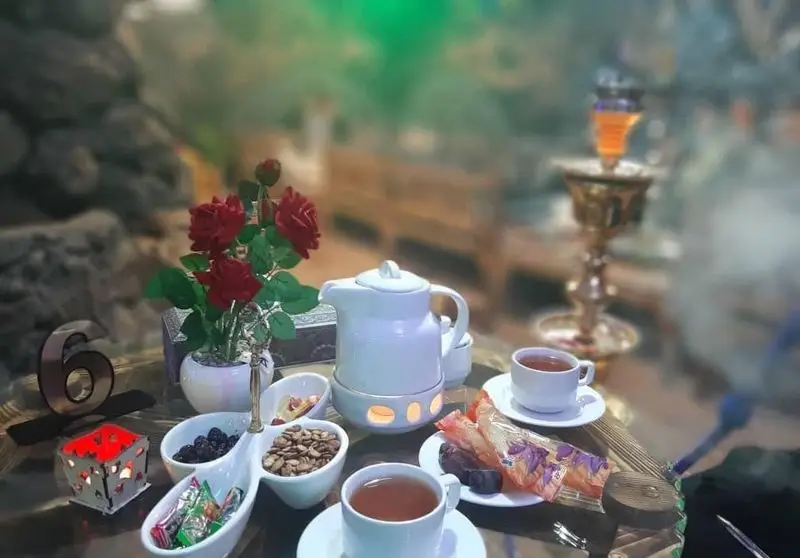 میز چای و تنقلات در سفره خانه سنتی ماندگار، با قوری، فنجان چای، گل رز و قلیان در فضایی سنتی و دلنشین