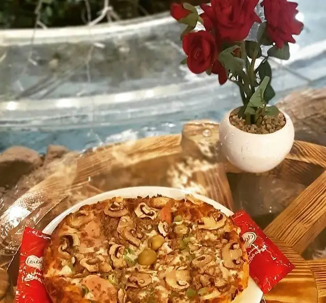 پیتزا قارچ و زیتون لذیذ در سفره خانه سنتی ماندگار. میز چوبی با گلدان گل رز قرمز و سس گوجه فرنگی، فضایی عالی برای تجربه غذای سنتی و مدرن.