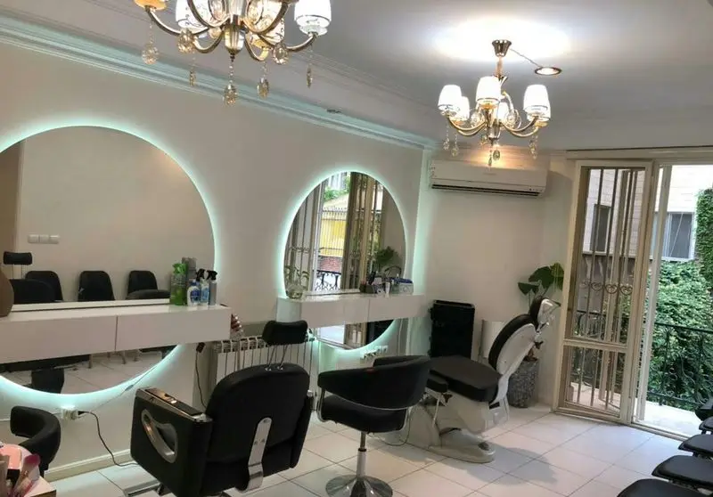 طراحی دکوراسیون داخلی آرایشگاه زنانه با آینههای بزرگ دایرهای بکلایت، صندلیهای اصلاح مدرن و لوسترهای شیک کریستالی