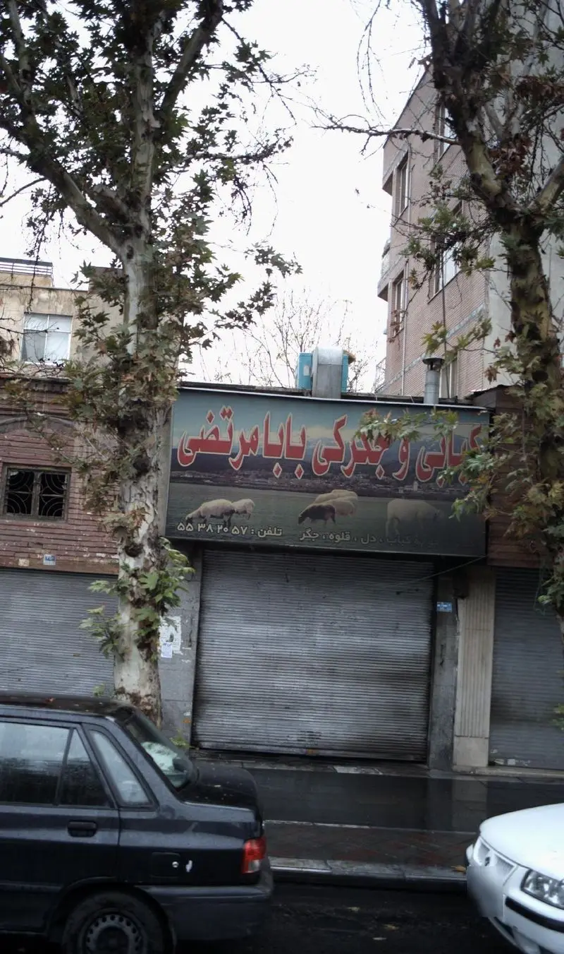 تصویر نمای خیابانی از کبابی و جگرکی بابا مرتضی با تابلوی حاوی عکس گوسفند و ذکر دل، قلوه، جگر و کباب. مغازه با کرکره فلزی بسته، در کنار درختان و خودروها.