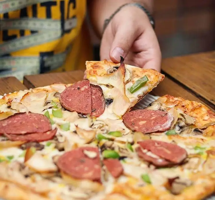 تصویری از یک پیتزای دلپذیر و اشتها آور با تاپینگ قارچ، کالباس و فلفل سبز در فودکورت رومانا که یک دست در حال برداشتن برش است.