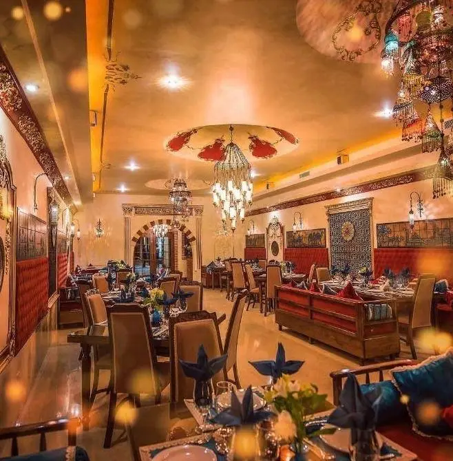 محیط داخلی مجلل کافه رستوران الحریرا با طراحی سنتی و دکوراسیون شرقی، لوسترهای خیره کننده، میز و صندلی‌های آماده برای پذیرایی با نورپردازی گرم.