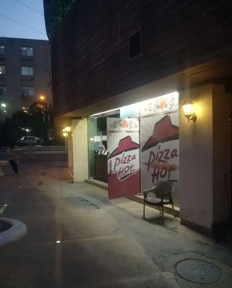 تصویری شبانه از شعبه پیتزا هات در شهرک غرب تهران، با لوگوی قرمز، ورودی روشن و صندلی در پیاده‌رو، آماده پذیرایی.