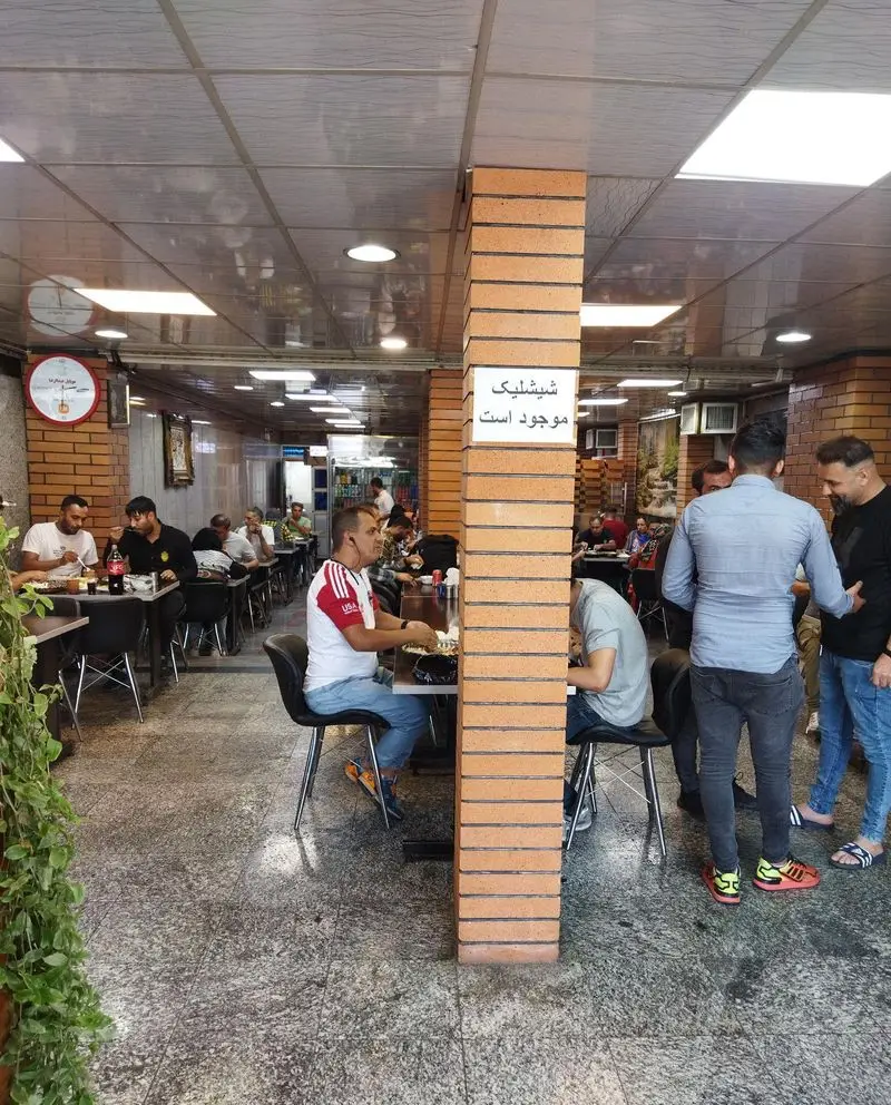 فضای داخلی رستوران حاتم با مشتریان در حال صرف غذا و تابلوی "شیشلیک موجود است" روی ستون آجری. محیطی پر جنب و جوش.