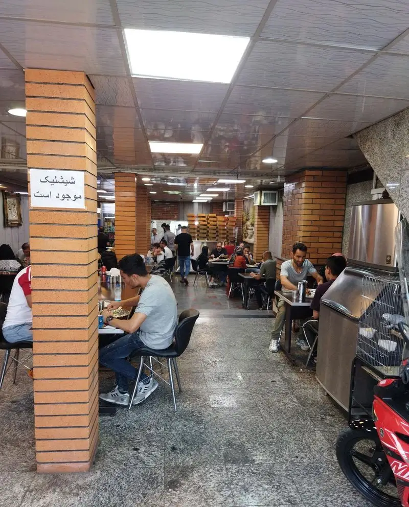 فضای داخلی دلپذیر رستوران حاتم با مشتریان در حال صرف غذا، میز و صندلی‌های راحت و ستون‌های آجری. شیشلیک نیز موجود است.