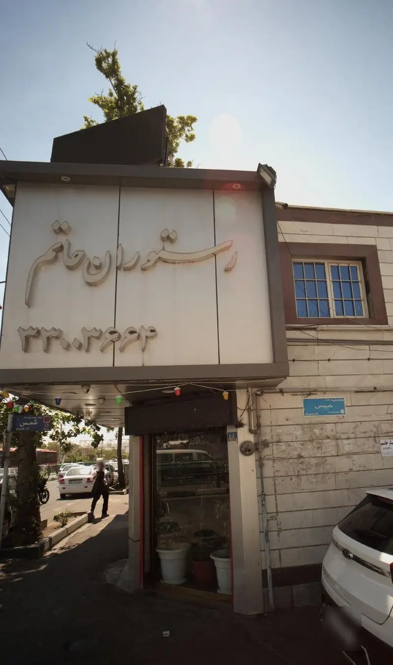 تصویر نمای بیرونی رستوران حاتم با تابلوی بزرگ و مشخصات شامل نام "رستوران حاتم" و شماره تلفن ۳۳۰۳۹۶۴. خیابان شمس با ماشین‌ها و فضای شهری نیز پیداست.