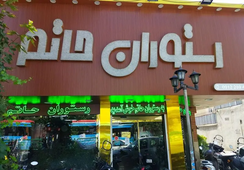 عکس سردر و نمای بیرونی رستوران حاتم در روز با تابلوی اصلی و نئونی، مکانی ایده‌آل برای غذای خوشمزه ایرانی و بیرون بر.