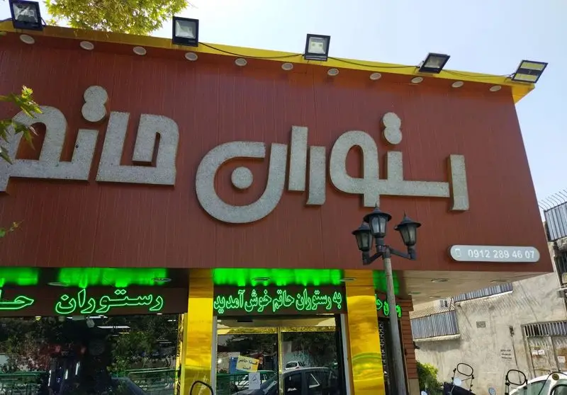 نمای اصلی رستوران حاتم با تابلوی برجسته نام، ورودی شیک و نورپردازی سبز. شامل شماره تماس مستقیم 09122894607 برای رزرو و اطلاعات بیشتر.