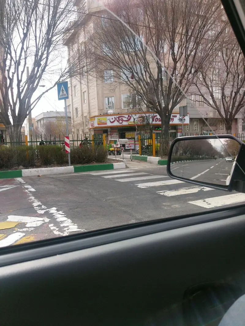 نمای بیرونی رستوران فست فود پیتزا توکان در یک روز آفتابی، واقع در کنار خیابانی شلوغ در فضای شهری، با درختان بدون برگ و خط‌کشی عابر پیاده.