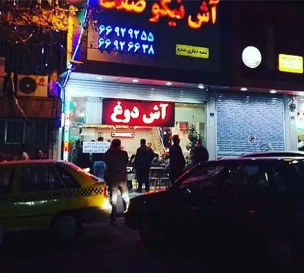تصویری از نمای بیرونی آش و حلیم پزی آش نیکوصفت در شب با تابلوی نئون و مشتریانی که آش دوغ می‌خرند و خودروها.