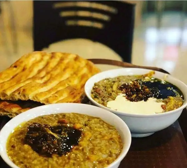 تصویر دو کاسه آش سنتی ایرانی با تزئینات کشک و نعناداغ و پیازداغ به همراه نان سنگک داغ، آماده سرو در آش نیکوصفت.