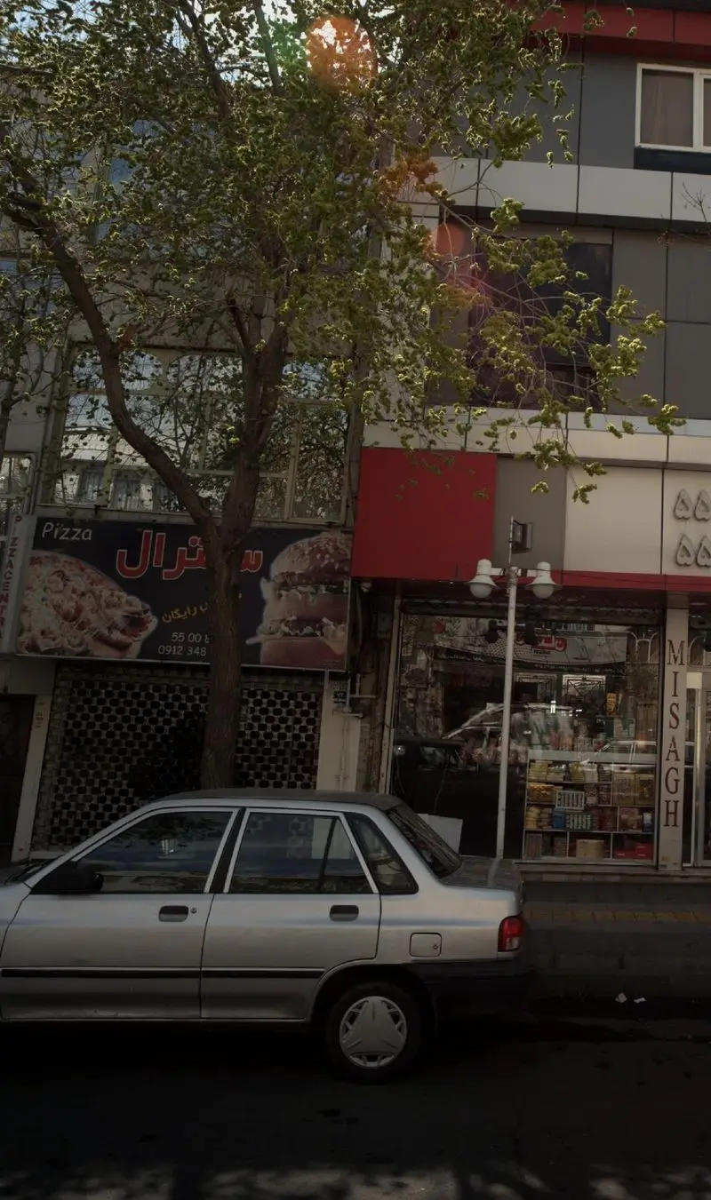 نمای خیابانی از فست فود پیتزا سنترال، تابلوی بزرگ با تصویر پیتزا و همبرگر دیده می‌شود، همراه با درختان و خودروی نقره‌ای پارک شده.