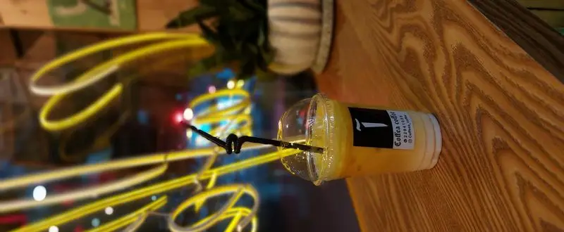 لیوان نوشیدنی خنک و خوشرنگ کافه آ (Coffea coffee) روی میز چوبی. لوگوی Coffea coffee و اطلاعات تماس. پس‌زمینه کافه با نورهای نئون زرد و رنگارنگ.