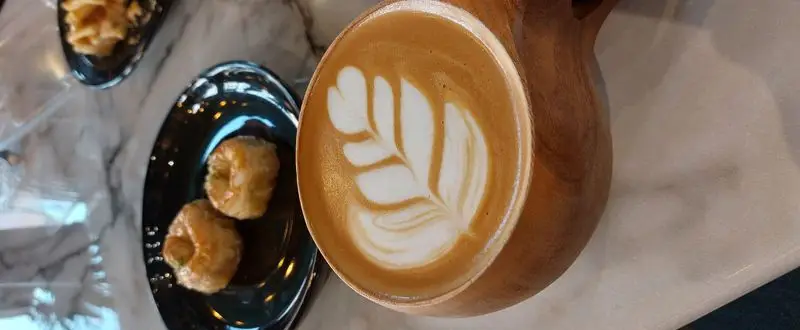 قهوه لاته آرت هنرمندانه در فنجان چوبی منحصربه‌فرد کافه آ، سرو شده با شیرینی‌های تازه و لذیذ برای لحظات آرامش‌بخش شما.